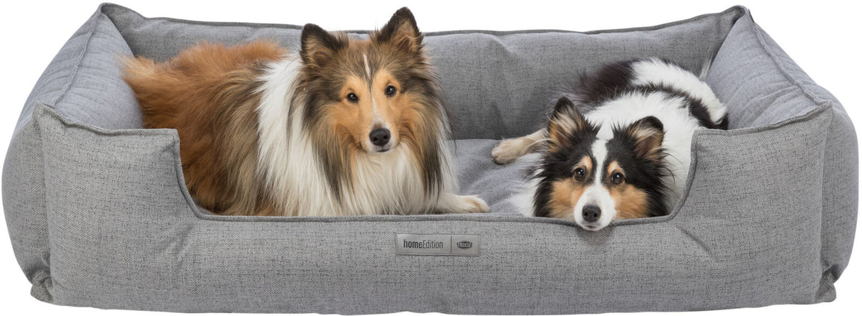 Zwei Hunde liegen entspannt in einem grauen Hundebett mit einem Etikett, das "homeEdition TRIXIE" zeigt.