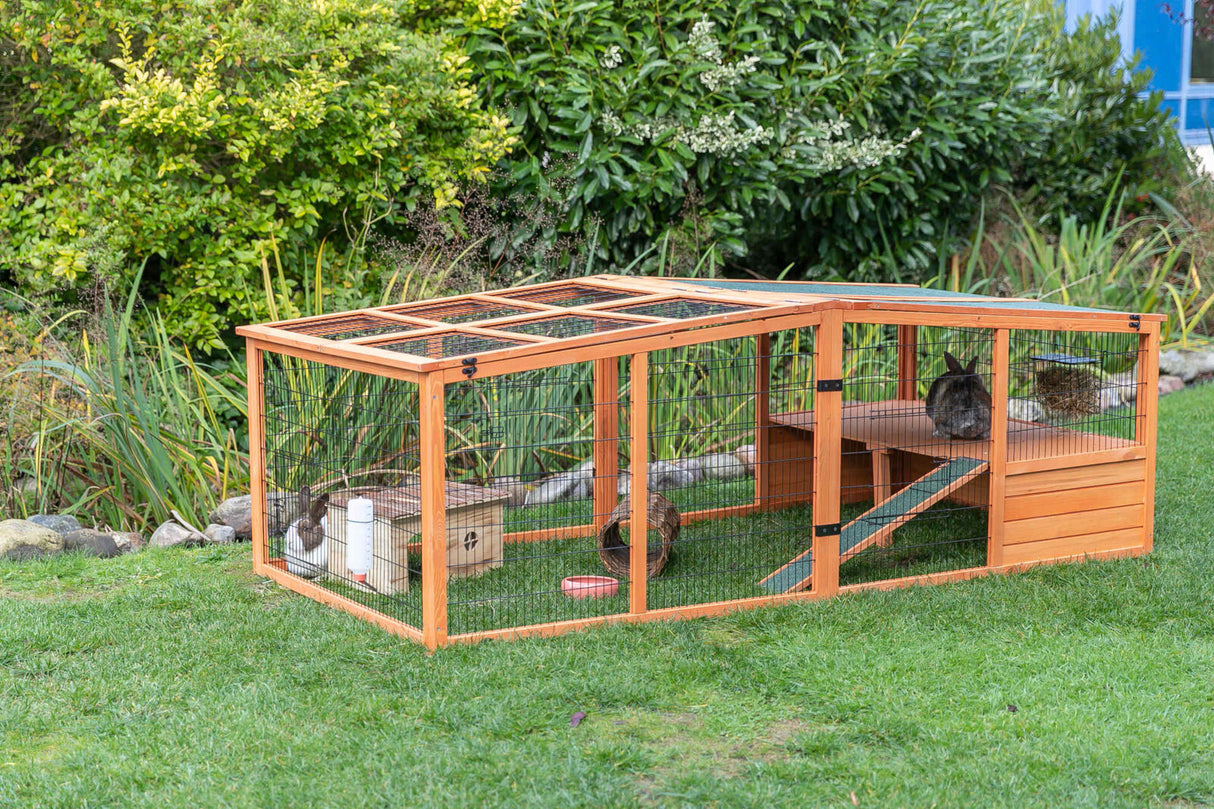 Holzgehege mit Gitterstruktur im Garten, enthält Kaninchen und Zubehör wie Futternapf und Häuschen.