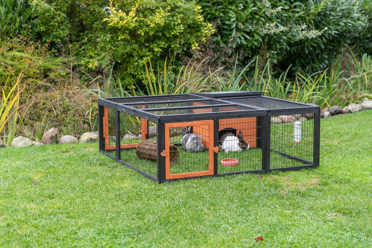 Kleintiergehege aus Metall mit Holzdetails auf grünem Rasen, innen zwei Kaninchen und Spielzeug.