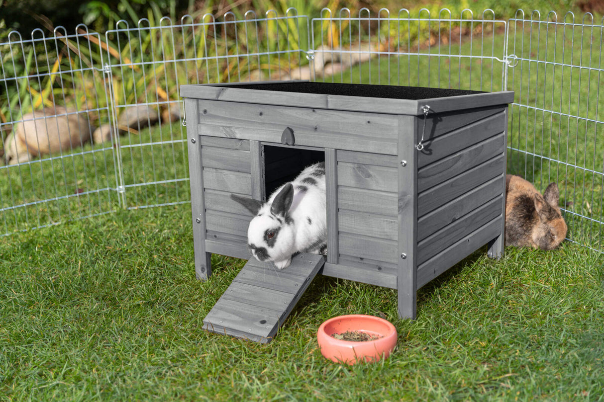 Grau gestrichener Holzstall mit geöffnetem Eingang, zwei Kaninchen im Freilaufgehege, Fressnapf im Vordergrund.