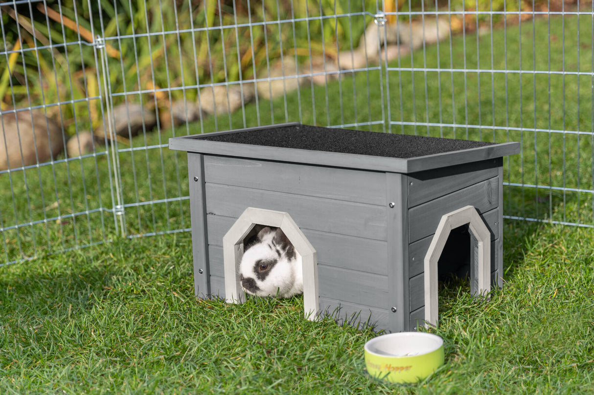 Graues Holzhäuschen in einem Drahtgehege auf Gras, ein Kaninchen schaut durch die Öffnung, daneben ein Napf.