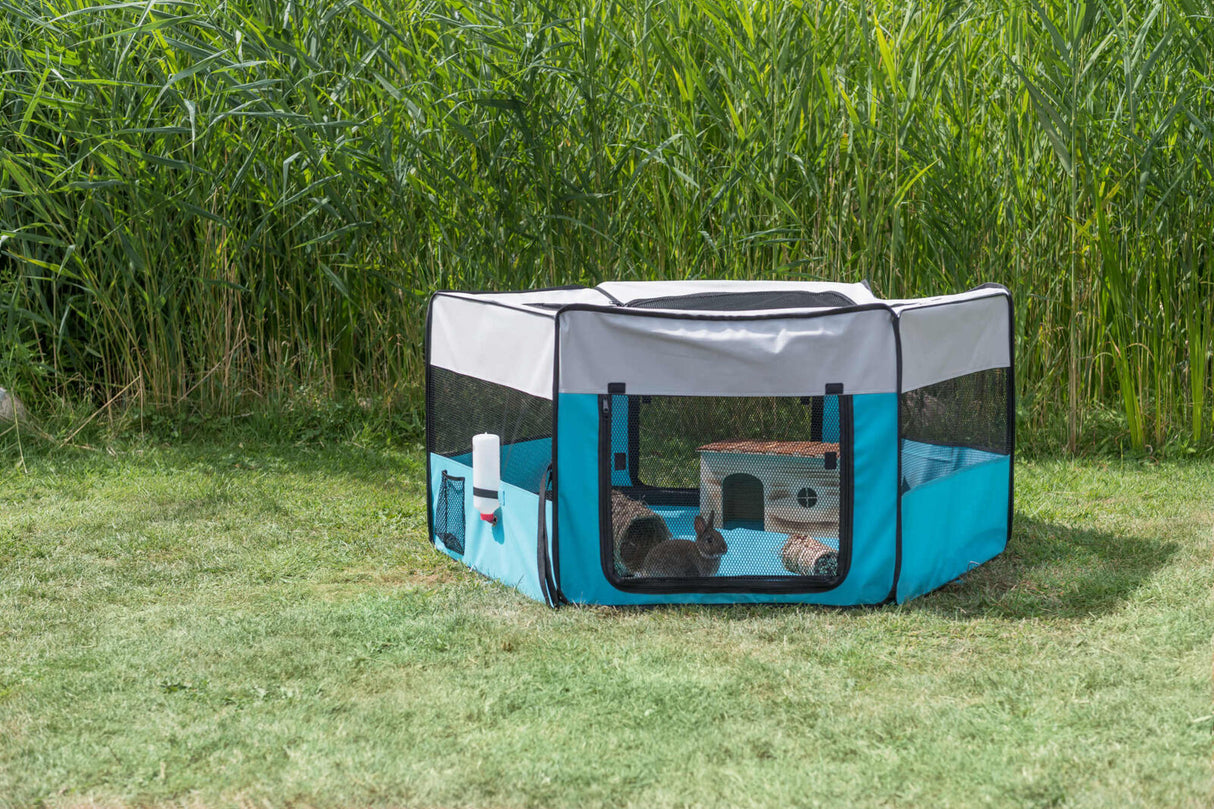 Ein blauer und grauer Laufstall mit Gitternetz steht auf einer Wiese neben hohen Gräsern, darin befinden sich zwei Kaninchen.