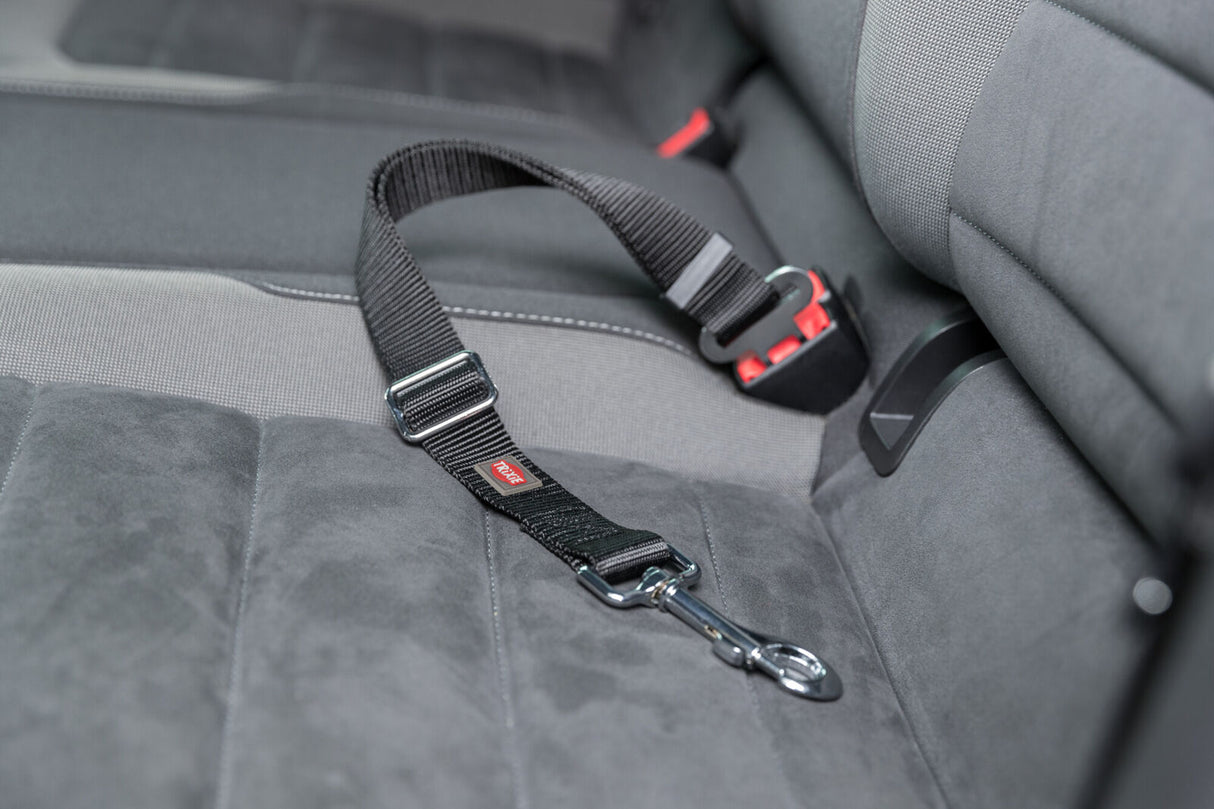 Ein schwarzer Sicherheitsgurt mit Metallklemme liegt auf einem grauen Autositz, sichtbar ist ein rotes Trageetikett.