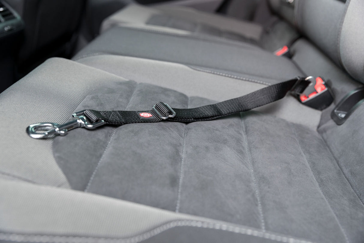 Schwarzer Sicherheitsgurt mit Karabiner auf grauem Autositz, Gurtende an rotem Verschluss befestigt.