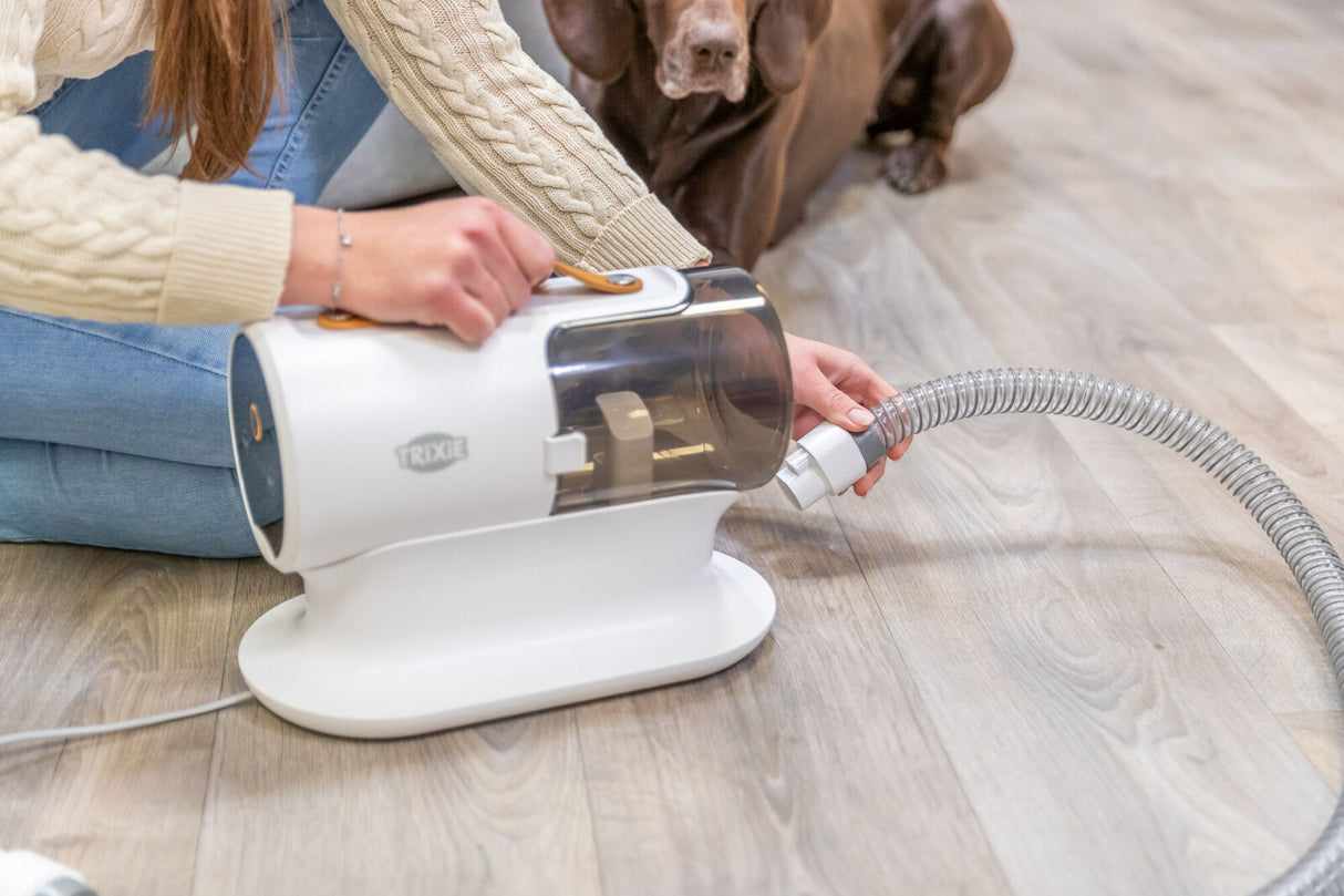Eine Person mit beige-grauem Pullover und blauer Jeans hält einen weißen Trixie-Staubsauger, während ein Hund daneben liegt.