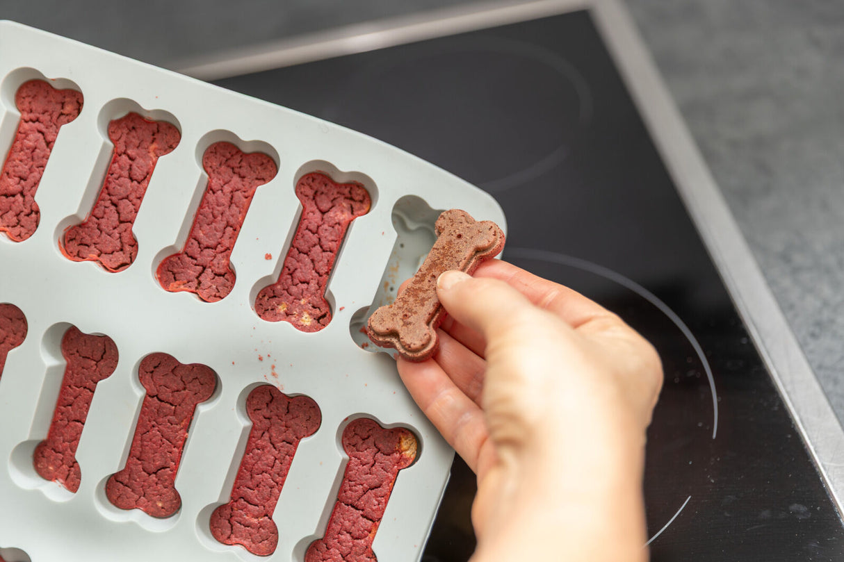 Hand hält knochenförmiges, rötliches Hundekeks aus einer Silikonform.