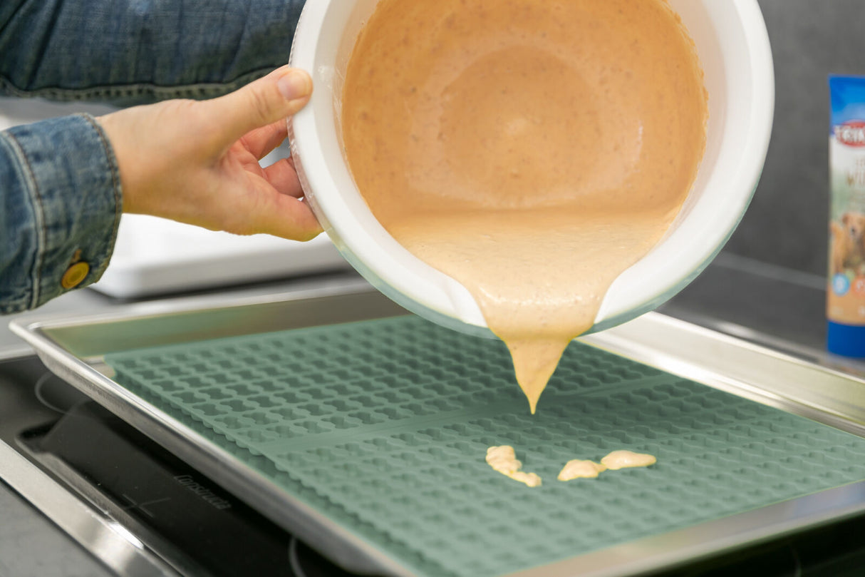 Person gießt braunen Teig aus einer Schüssel auf eine grüne Backmatte in einem Backblech auf einem Herd.