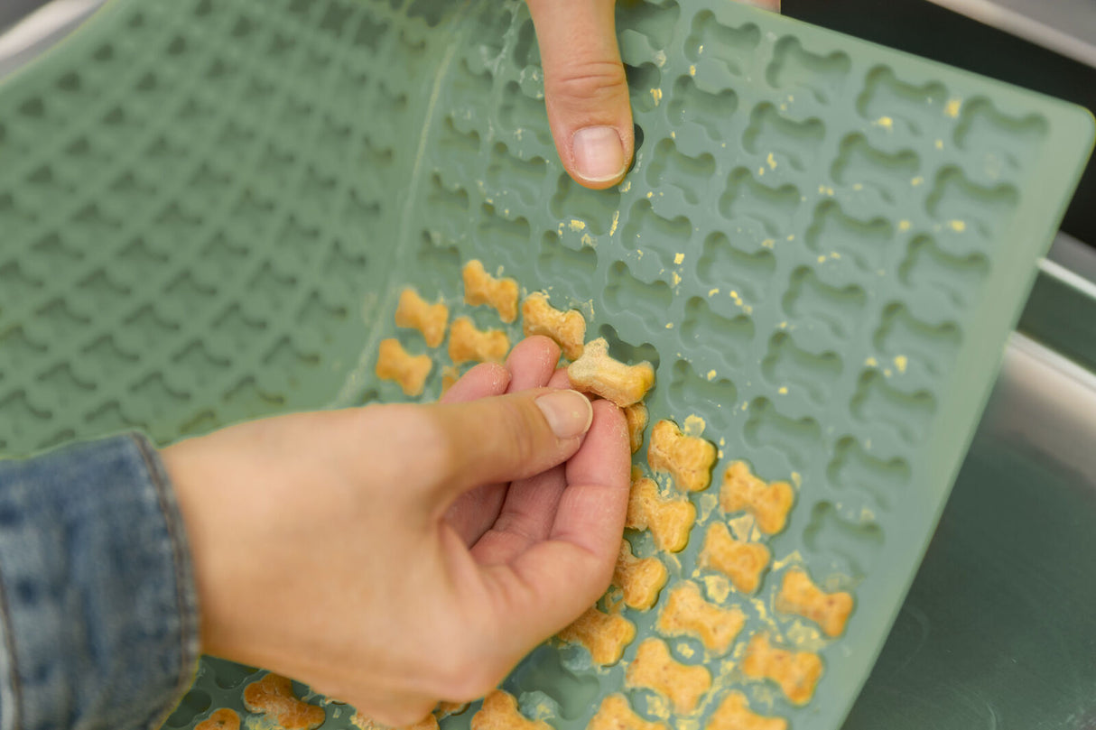 Eine Person entnimmt kleine, knochenförmige, orangefarbene Kekse aus einer grünen Silikonform.