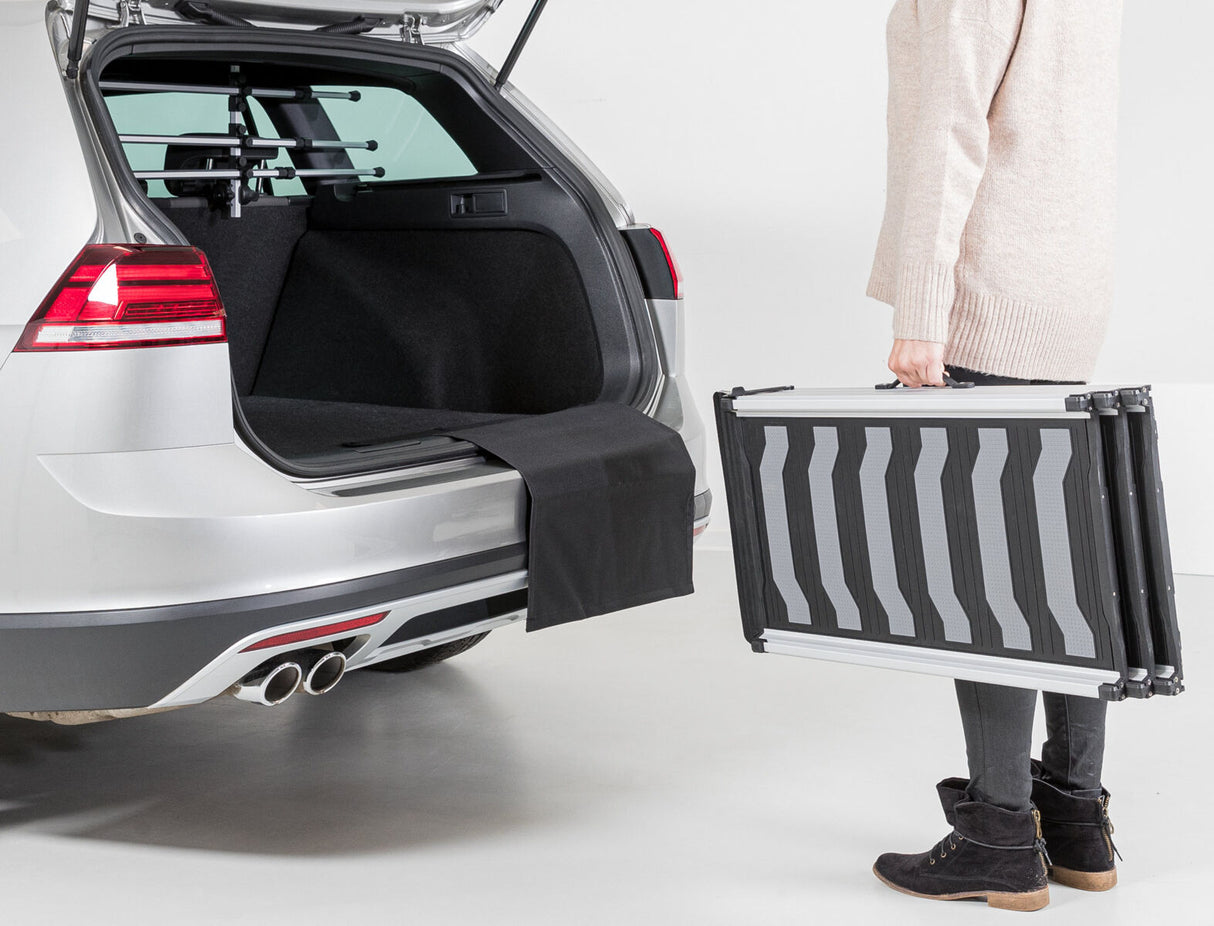 Eine Person steht neben einem geöffneten Kofferraum eines silbernen Autos und hält eine zusammenklappbare Rampe.