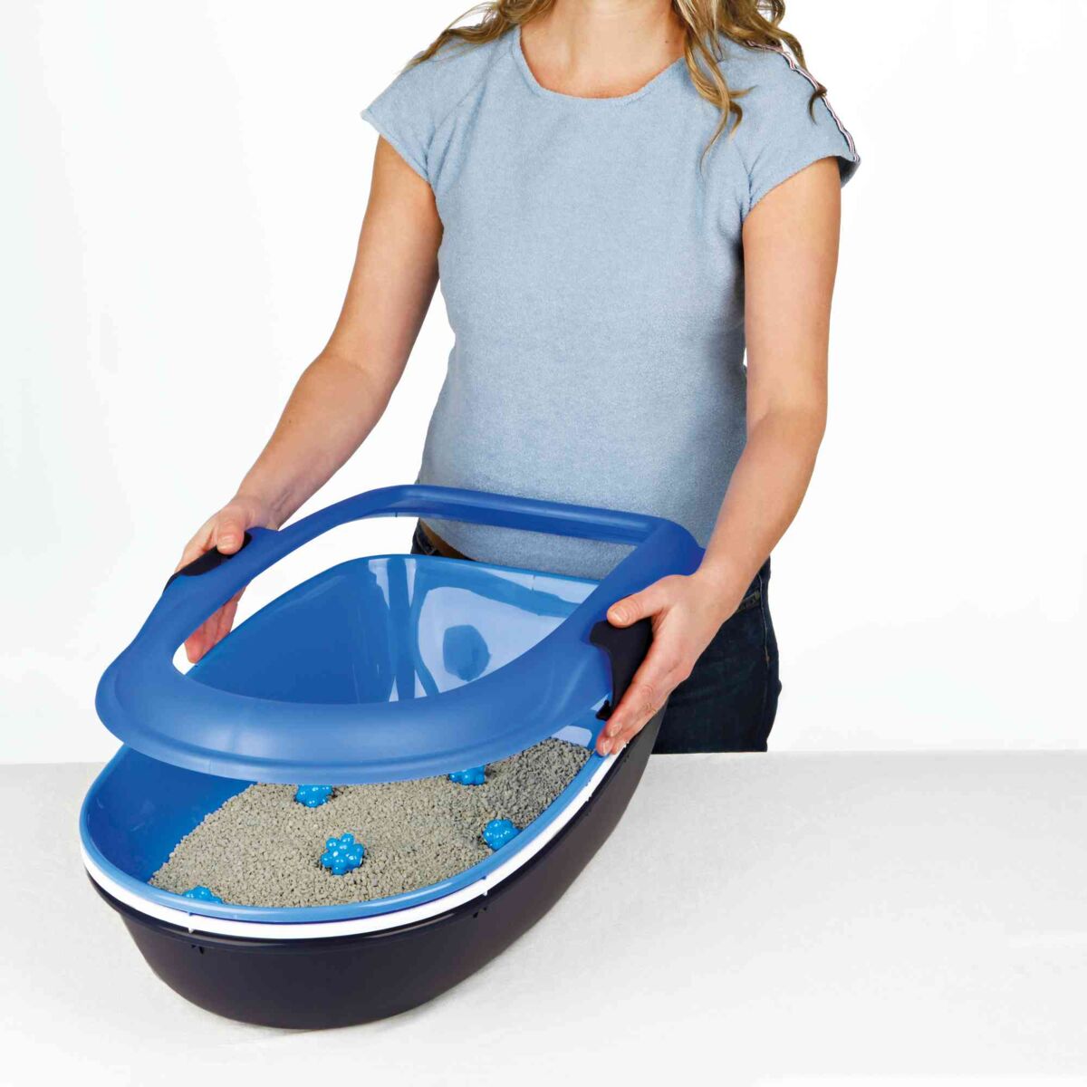 Eine Person in einem blauen Shirt hält einen blauen Deckel einer Katzentoilette mit Streu und blauen Elementen.