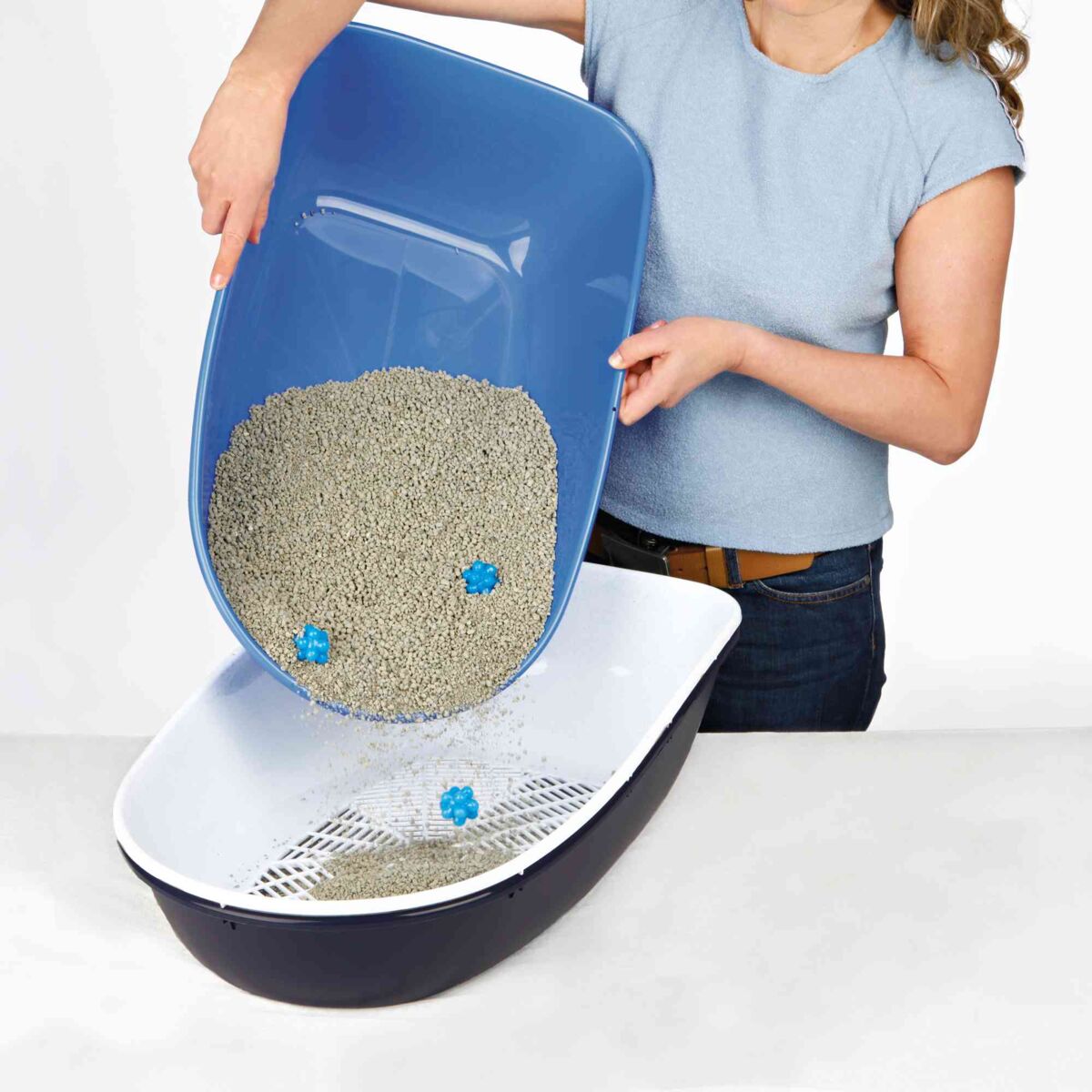 Eine Person in einem hellblauen Oberteil schüttet Katzenstreu aus einem blauen Behälter in eine schwarze Katzentoilette.