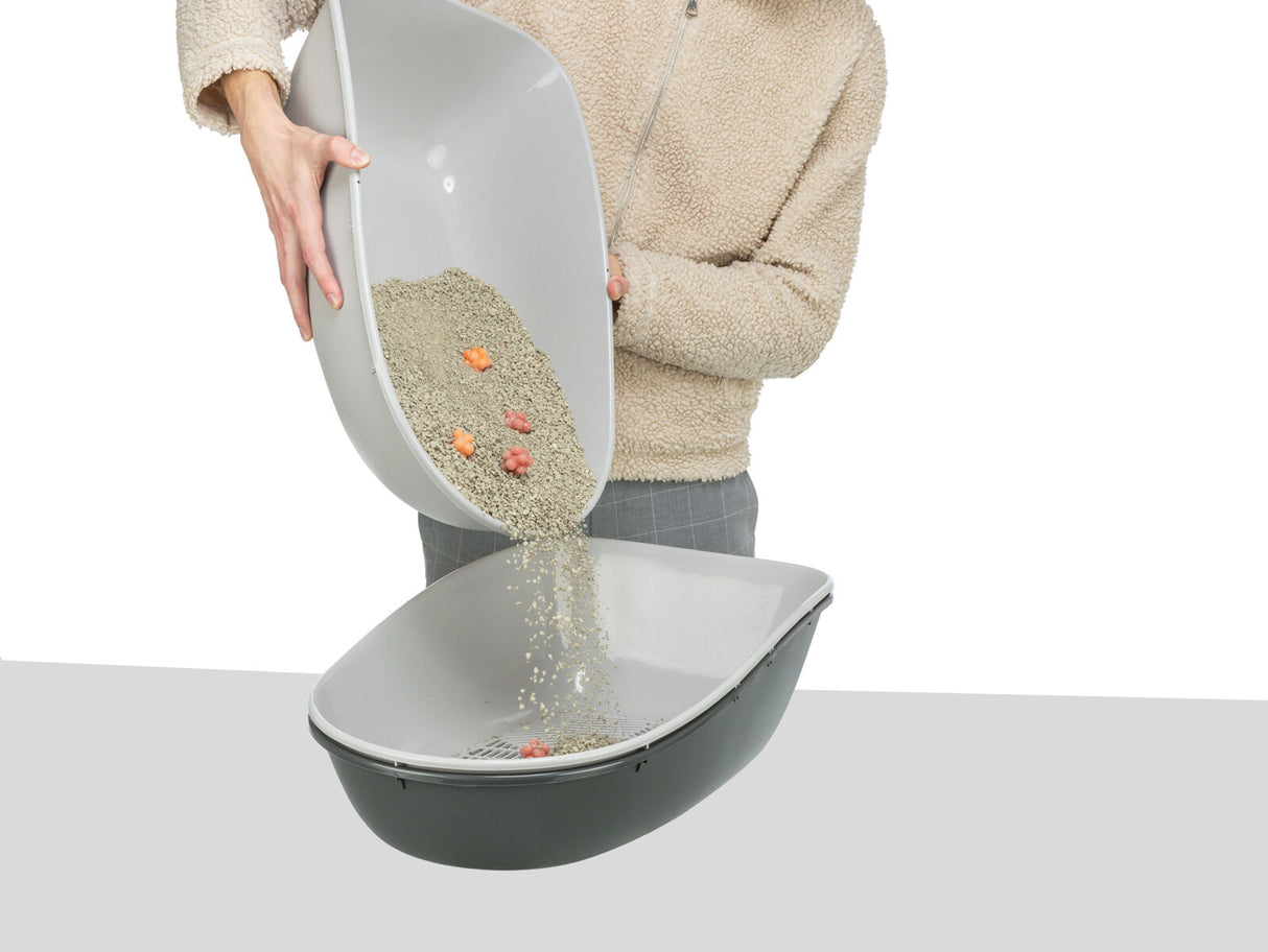 Person schüttet Katzenstreu von einer Schale in eine andere, beide hellgrau, auf grauem Hintergrund.