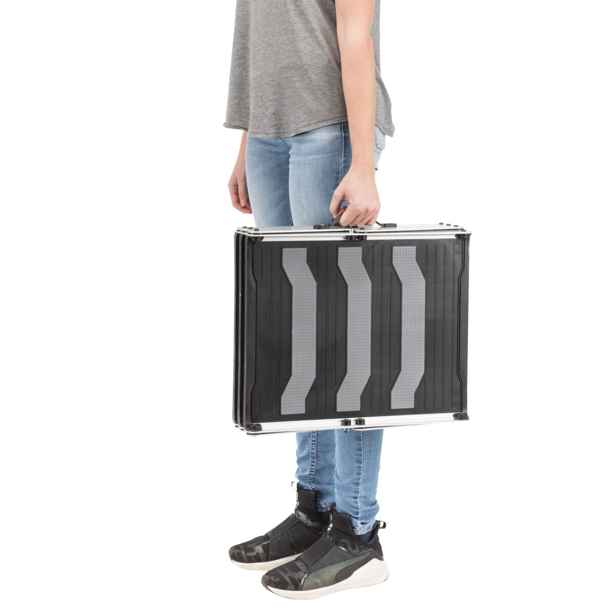 Person trägt einen schwarzen Koffer mit grauen Streifen seitlich, trägt graues Shirt, Jeans und schwarze Schuhe.