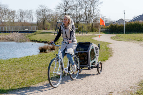 Eine Frau mit blonden langen Haaren fährt mit einem weißen Fahrrad auf einem Sandweg neben einem Teich. Sie zieht einen grauen Fahrradanhänger.