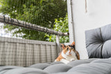 Dreifarbige Katze liegt auf gepolstertem Balkon mit Netz und Pflanzen im Hintergrund.