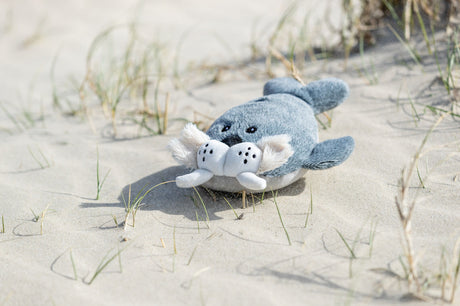 Ein graues Plüschtier in Form einer Robbe liegt auf Sand mit spärlichem Gras.