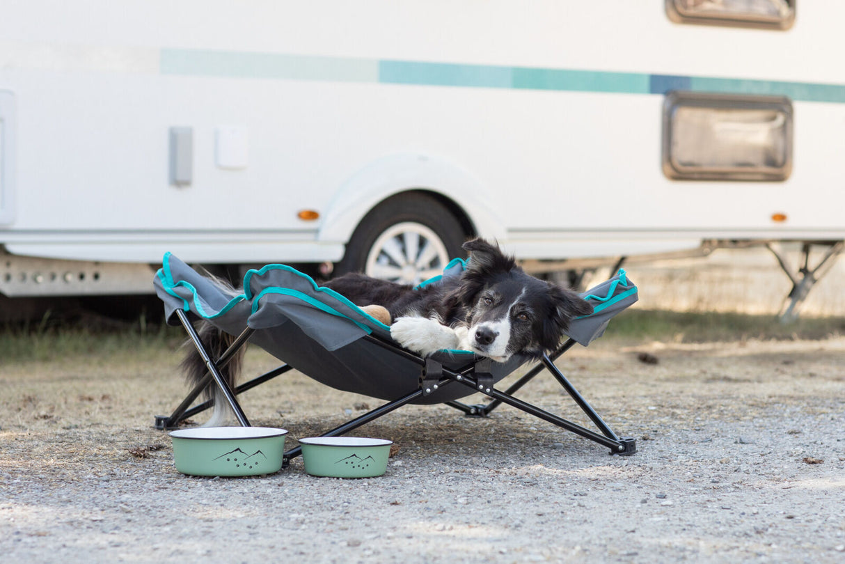Hund liegt entspannt in einem faltbaren Hundebett vor einem Wohnwagen auf Kiesboden.