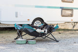 Hund liegt entspannt in einem faltbaren Hundebett vor einem Wohnwagen auf Kiesboden.