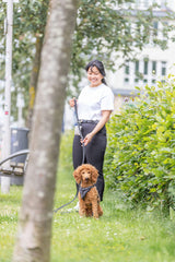 Frau mit weißem T-Shirt hält einen kleinen, lockigen Hund an der Leine auf einer grünen Wiese.
