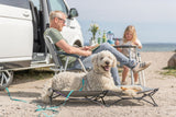 Mann und Kind sitzen neben Hund auf Hundebett vor einem weißen Camper am Strand.