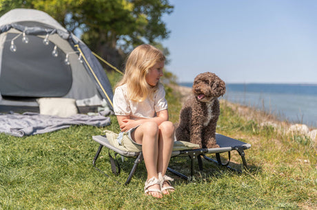 Mädchen sitzt neben einem braunen Hund vor einem Zelt am Seeufer.