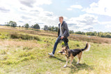 Mensch mit grauer Jacke geht mit großem Hund, der Spielzeug trägt, auf einer Wiese spazieren.