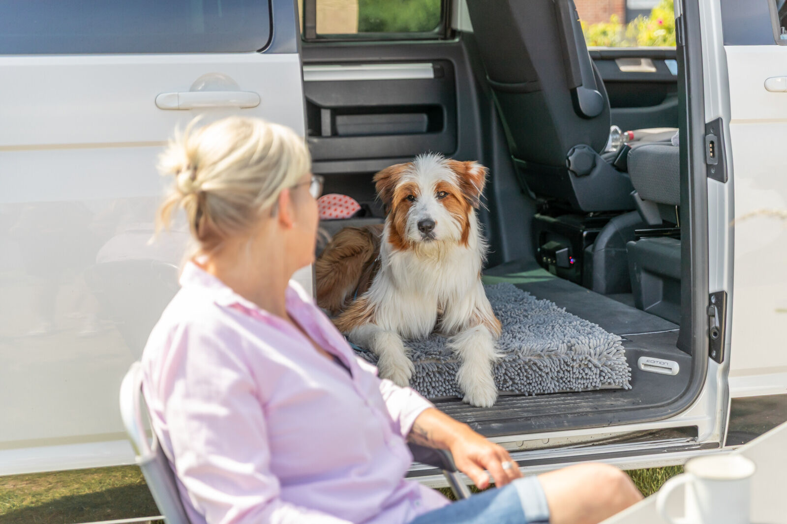 Hund liegt auf grauem Teppich im geöffneten Kofferraum eines Autos neben einer sitzenden Frau.