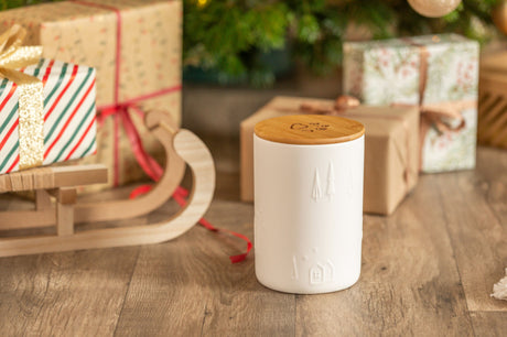 Weiße Dose mit Holzdeckel auf Holzboden neben Geschenken und einem Schlitten vor Tannenbaum.