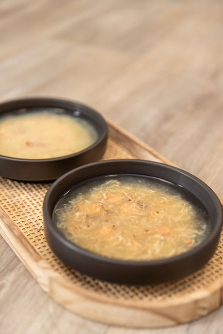 Zwei schwarze Schüsseln mit heller Suppe auf einem Tablett aus Holzgeflecht.