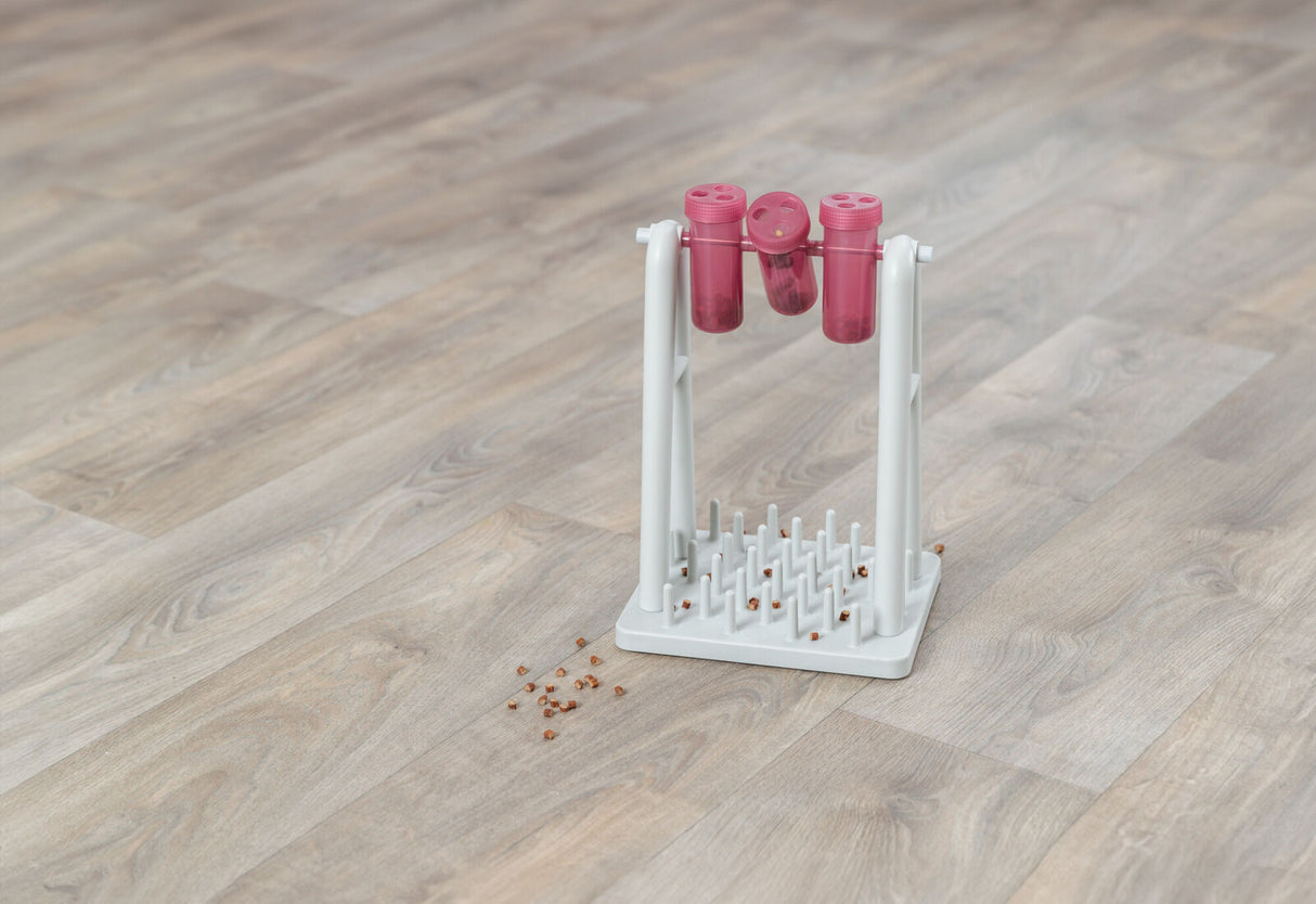 Weißer Ständer mit drei rosa Behältern und Stäben auf Holzboden, daneben verstreute braune Körner.