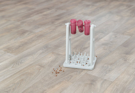 Weißer Ständer mit drei rosa Behältern und Stäben auf Holzboden, daneben verstreute braune Körner.