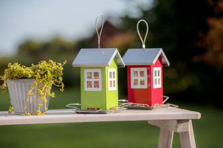 Zwei vogelhausförmige Futterspender aus Metall in Grün und Rot mit transparenten Fenstern auf Holztisch neben Pflanzentopf.