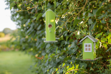 Zwei grüne Vogelfutterspender in unterschiedlichen Formen hängen an Ästen vor einem Hintergrund aus dichten, grünen Blättern.