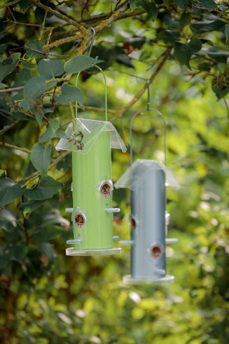 Zwei hängende Vogelfutterspender, einer in Grün und der andere in Blau, umgeben von grünen Blättern.