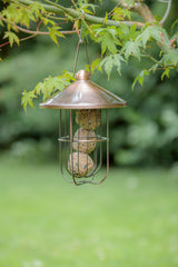 Metallener Vogel-Futterspender in Käfigform hängt an einem Ast mit grünen Blättern.