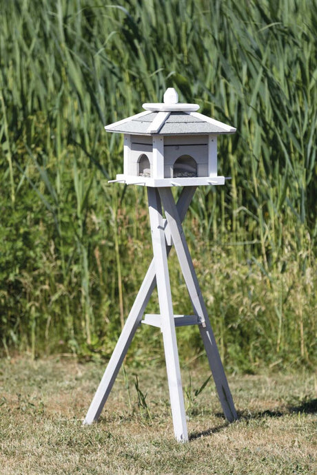 Weißes Vogelhaus auf hohem Ständer vor grüner Pflanzenkulisse.