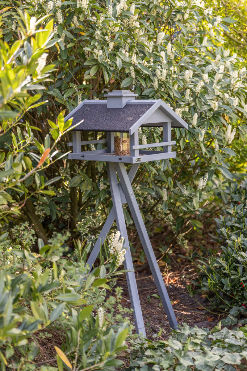 Ein graues Vogelhäuschen mit schwarzem Dach steht auf einem dreibeinigen Ständer vor dichtem grünem Laub.