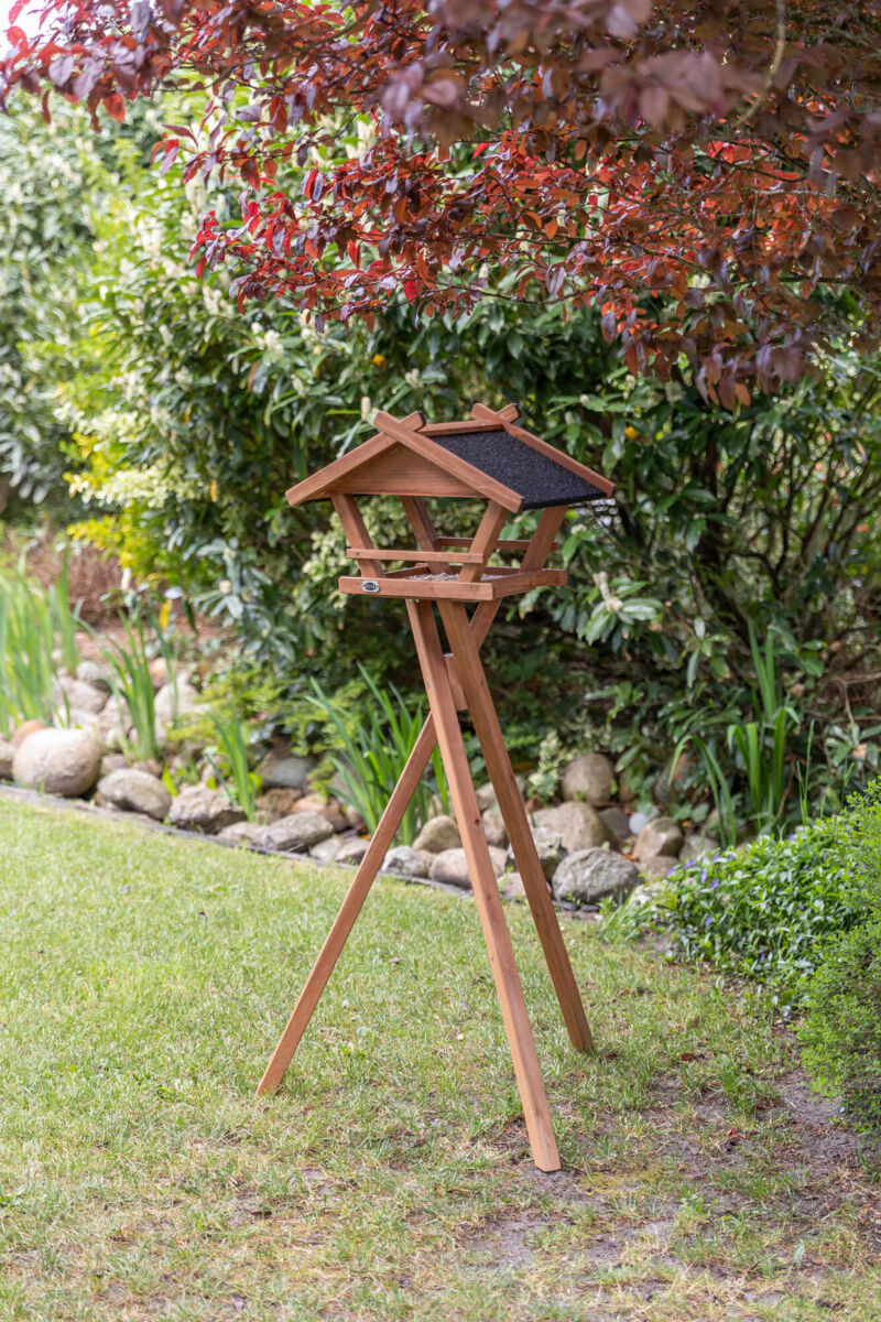 Holz-Vogelhaus mit schwarzem Dach auf dreibeinigem Ständer, umgeben von grünem Garten und rotem Laub.