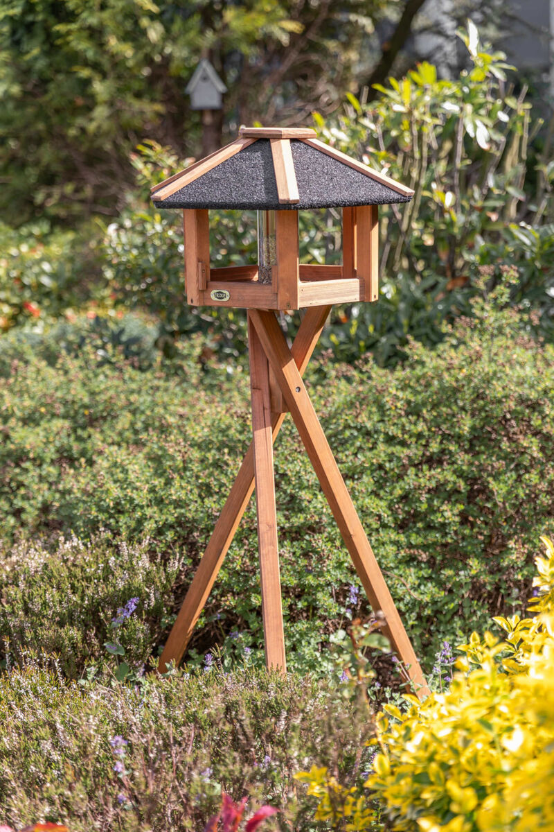 Ein braunes, holzernes Vogelhaus mit schwarzem Dach steht auf einem dreibeinigen Gestell im grünen Garten.