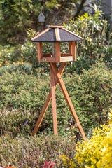 Ein braunes, holzernes Vogelhaus mit schwarzem Dach steht auf einem dreibeinigen Gestell im grünen Garten.