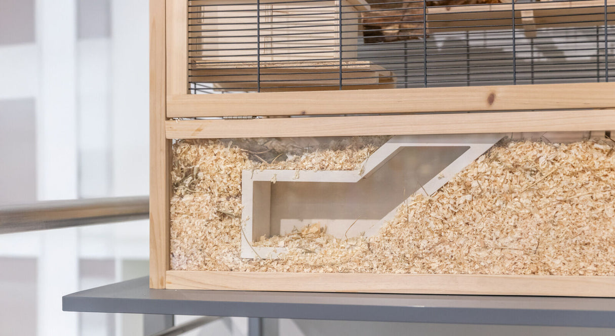 Holzgehege mit Gitter, teilweise gefüllt mit Holzspänen, auf einer grauen Oberfläche stehend.