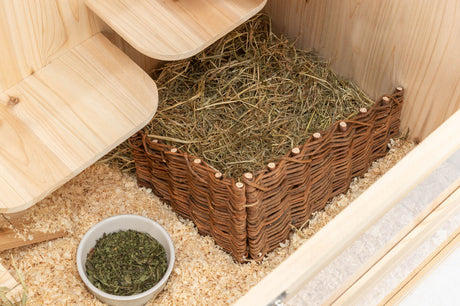 Ein Holzkäfig mit Heu, geflochtenem Korb und Schalen mit grünem Futter auf Streu.