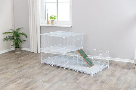 Ein weißes Gitterkäfig mit zwei Ebenen und einer Rampe steht auf einem Holzboden in einem hellen Raum.