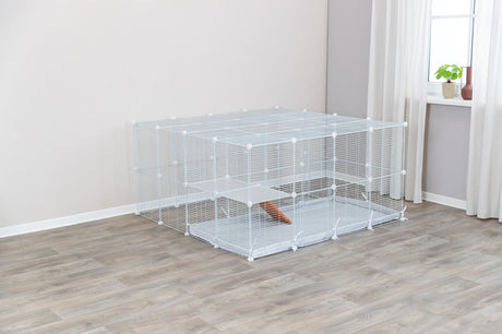 Ein rechteckiger, weißer Gitterkäfig mit mehreren Ebenen steht auf einem Holzboden in einem hellen Raum mit Fenster.