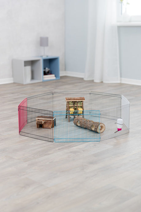 Ein Metallgehege mit bunten Elementen auf Boden, darin ein Holzhaus, Tunnel und eine Wasserflasche.