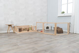 Holzgehege mit Drahtgitter und Einstreu auf hellem Holzboden, links Tisch mit Glasflasche, rechts Fenster mit Vorhang.