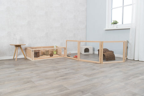 Holzgehege mit Drahtgitter und Einstreu auf hellem Holzboden, links Tisch mit Glasflasche, rechts Fenster mit Vorhang.