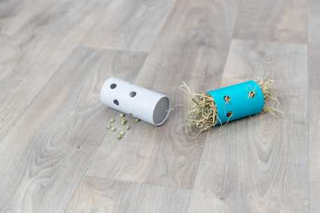 Zwei zylinderförmige Objekte mit Löchern, eines mit grünem Heu, das andere mit verstreuten Erbsen auf Holzfußboden.