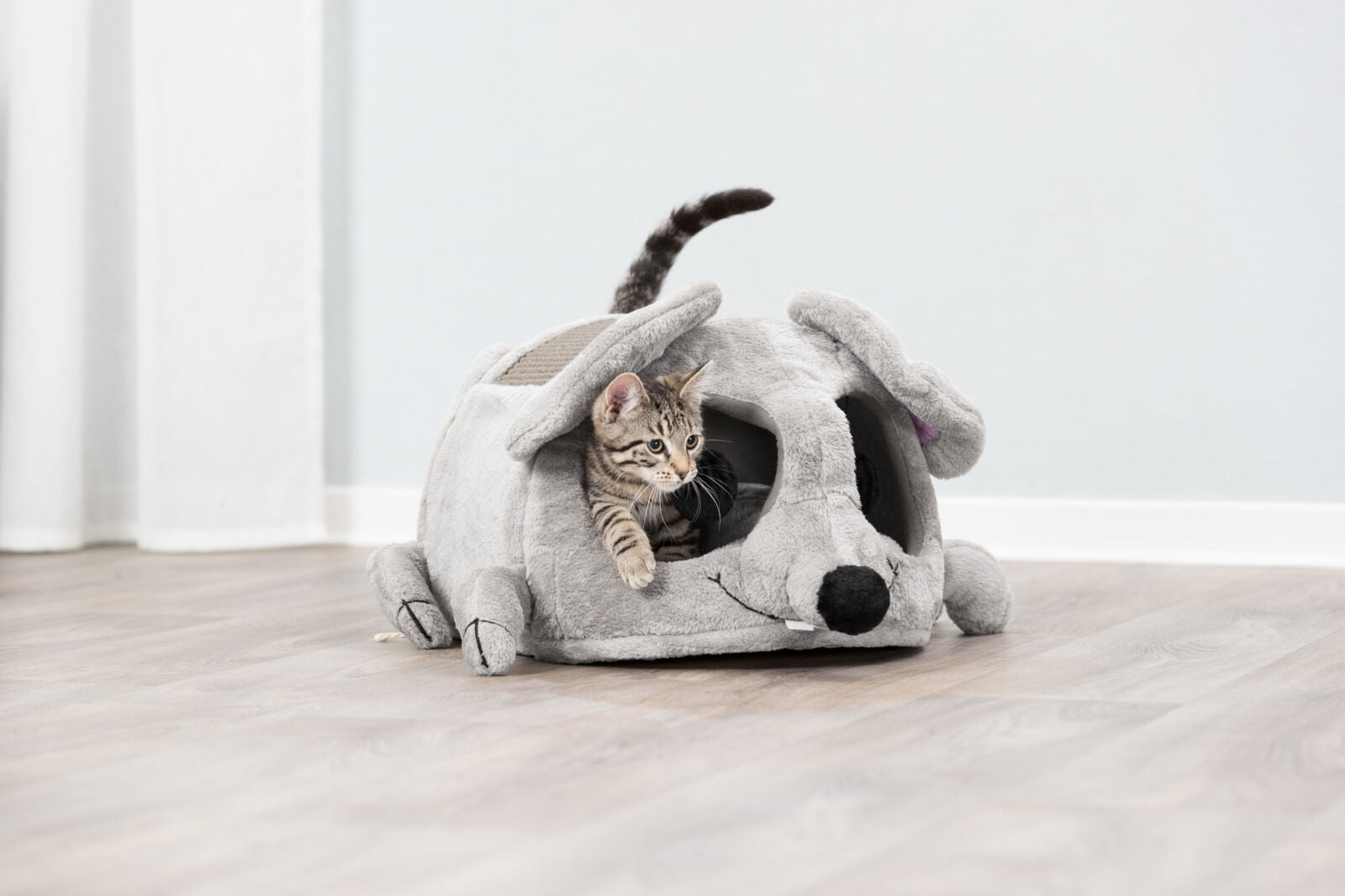 Kleine Katze schaut aus einer grauen Plüschhöhle in Form einer Maus auf hellem Holzboden hervor.