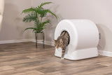 Eine getigerte Katze verlässt eine weiße, runde Katzentoilette mit einer Pflanze im Hintergrund auf einem Holzboden.